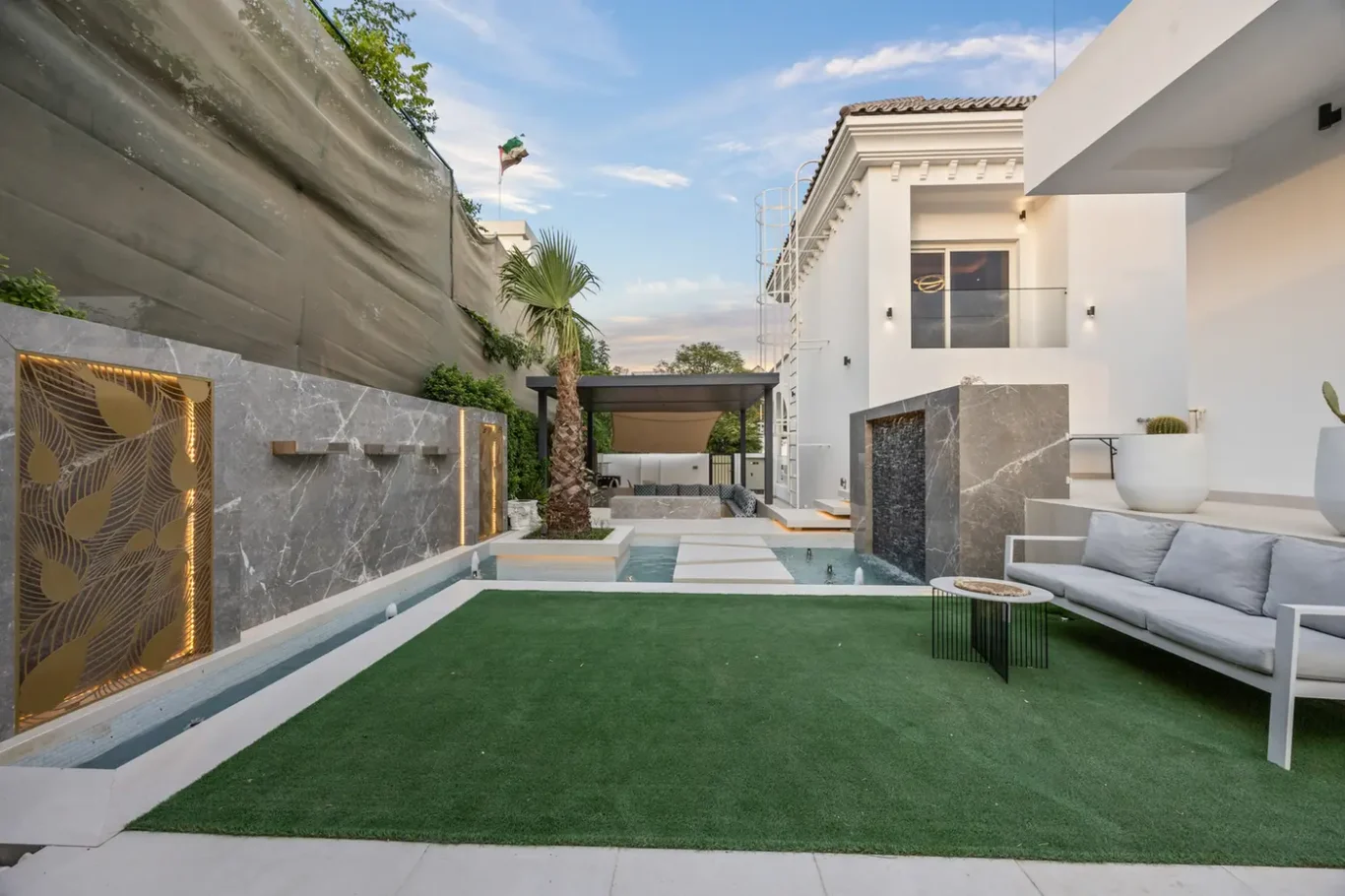 Modern white-house backyard with a rectangular pool, artificial turf, palm tree, and gray marble walls with backlit panels on a sunny day