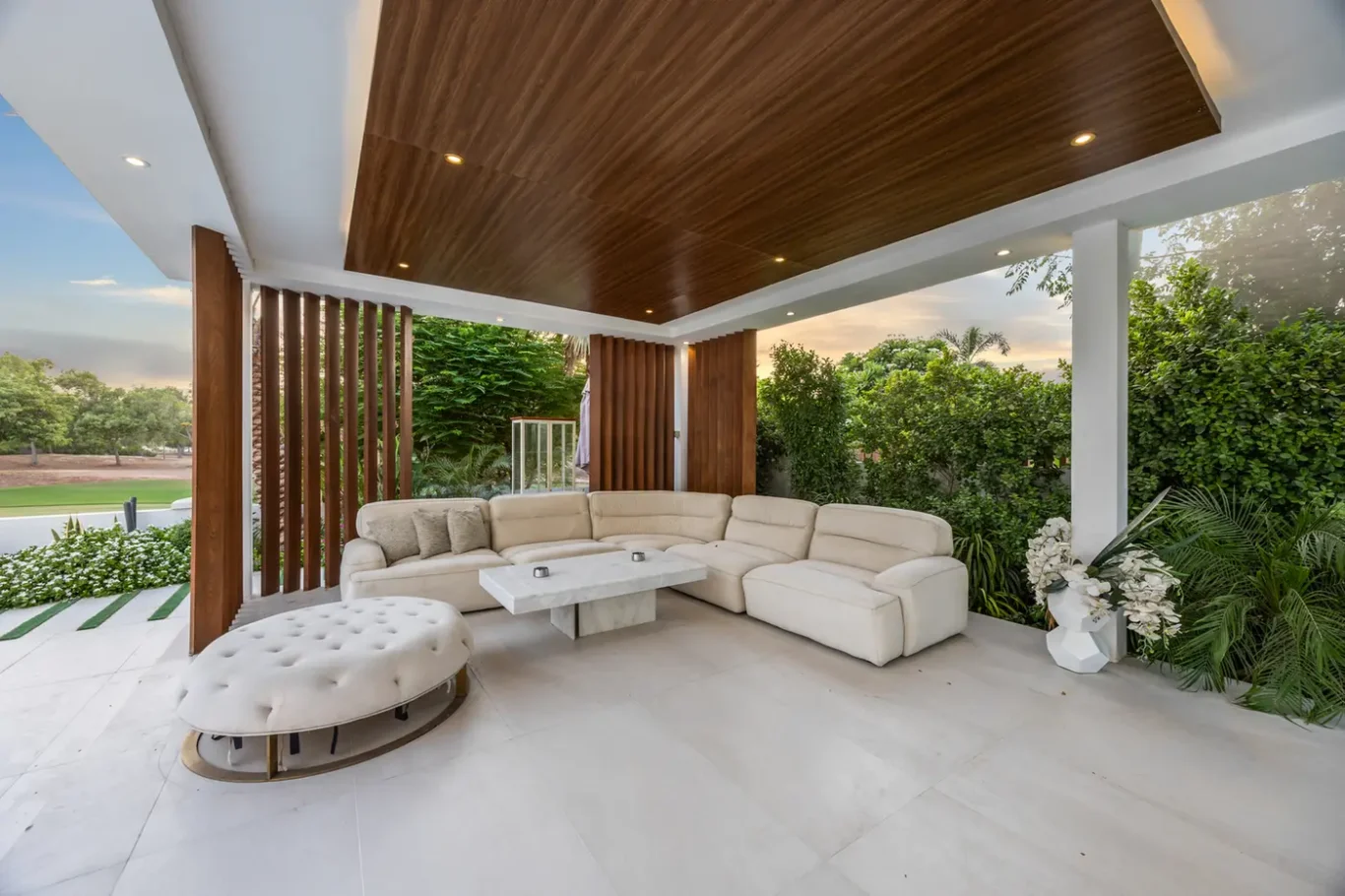 Covered outdoor seating area with a large cream sectional sofa, white tufted ottoman, and wooden ceiling panels amid greenery outside a modern home driveway.