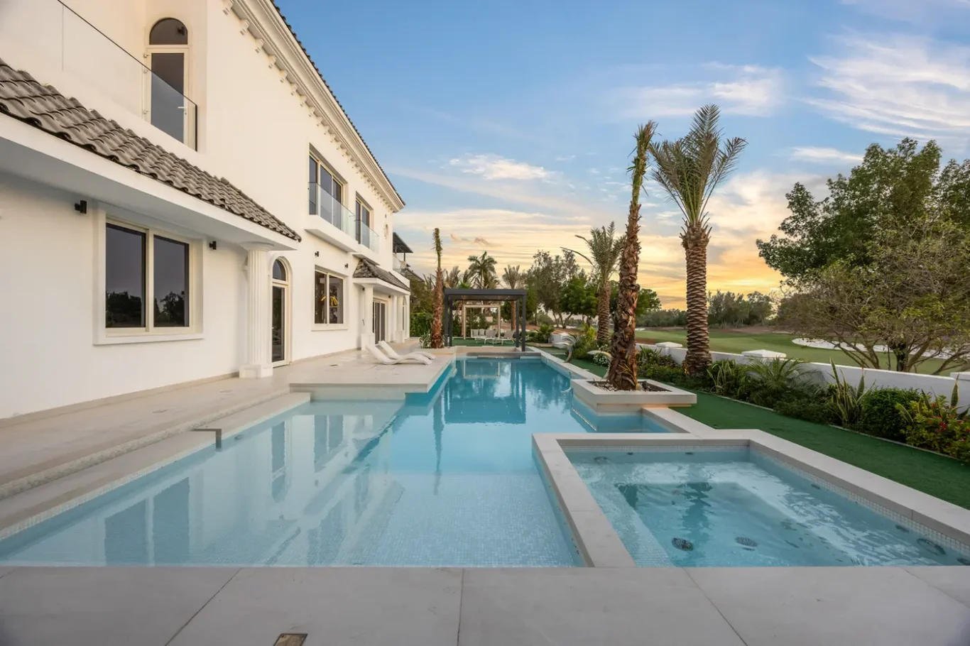 White modern villa with a long rectangular pool and spa, palm trees, and a sunset sky in the background.