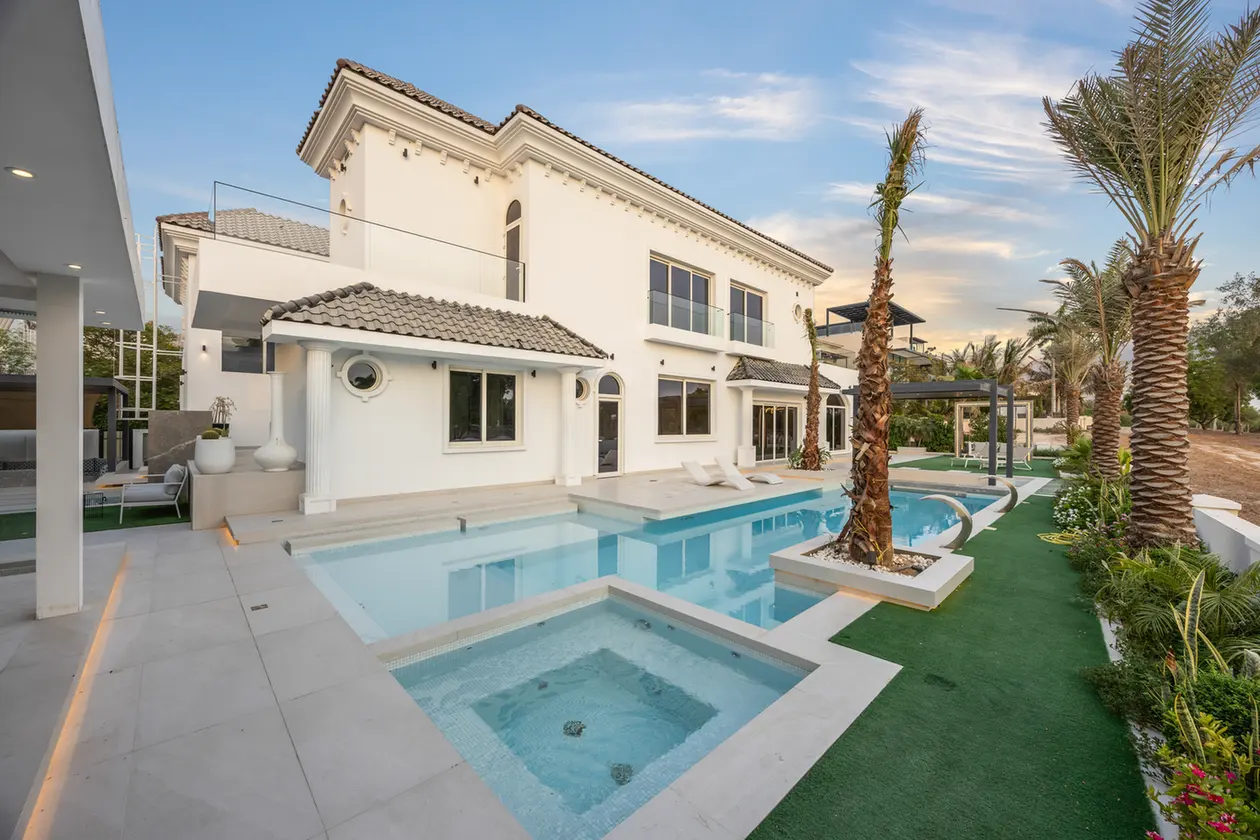White two-story villa with a rectangular pool and spa, palm trees, and lounge chairs on a sunny day.