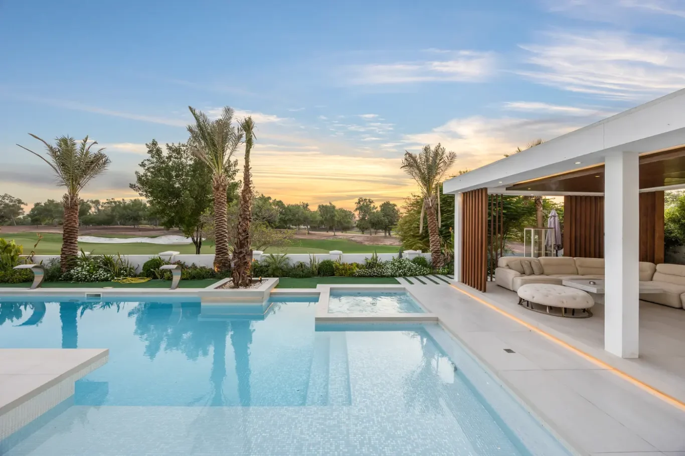 Luxurious backyard with an in-ground pool, spa, and a covered seating area overlooking a green lawn and palm trees at sunset .