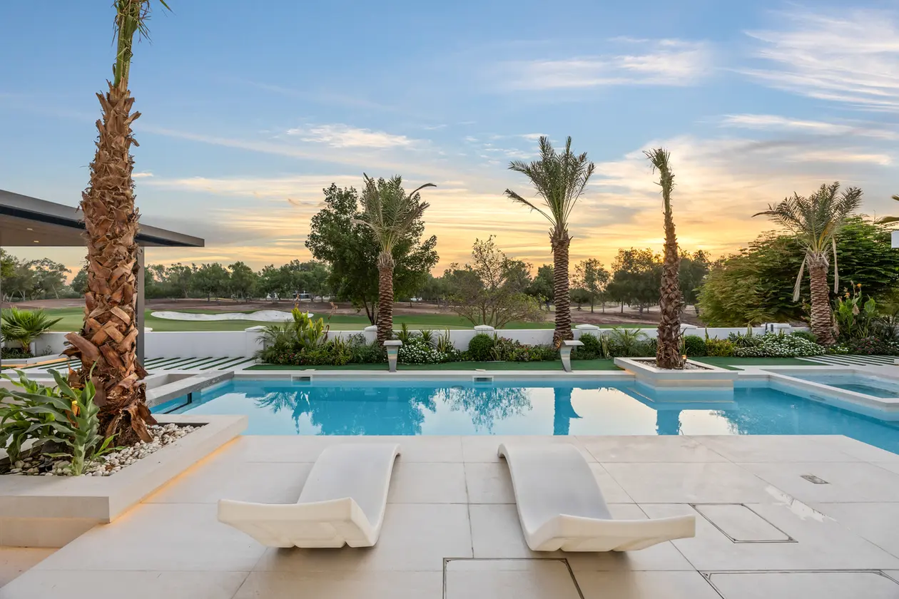 Luxurious poolside area at sunset with two white curved lounge chairs, palm trees, and landscaped greenery surrounding a blue pool.