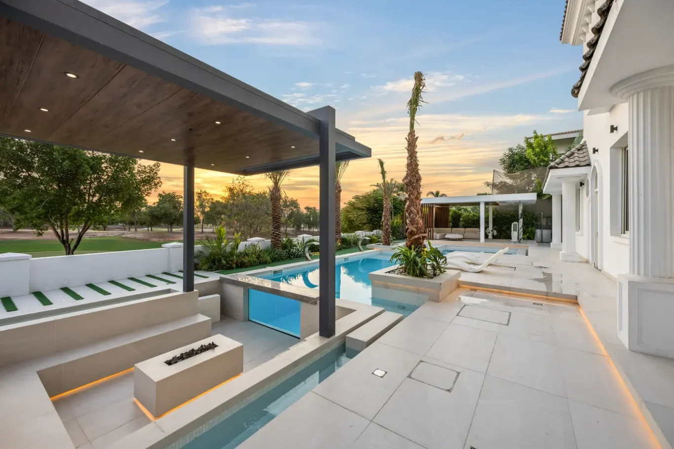 Modern backyard pool area at sunset with a white villa, a rectangular pool, palm trees, and a covered seating area.