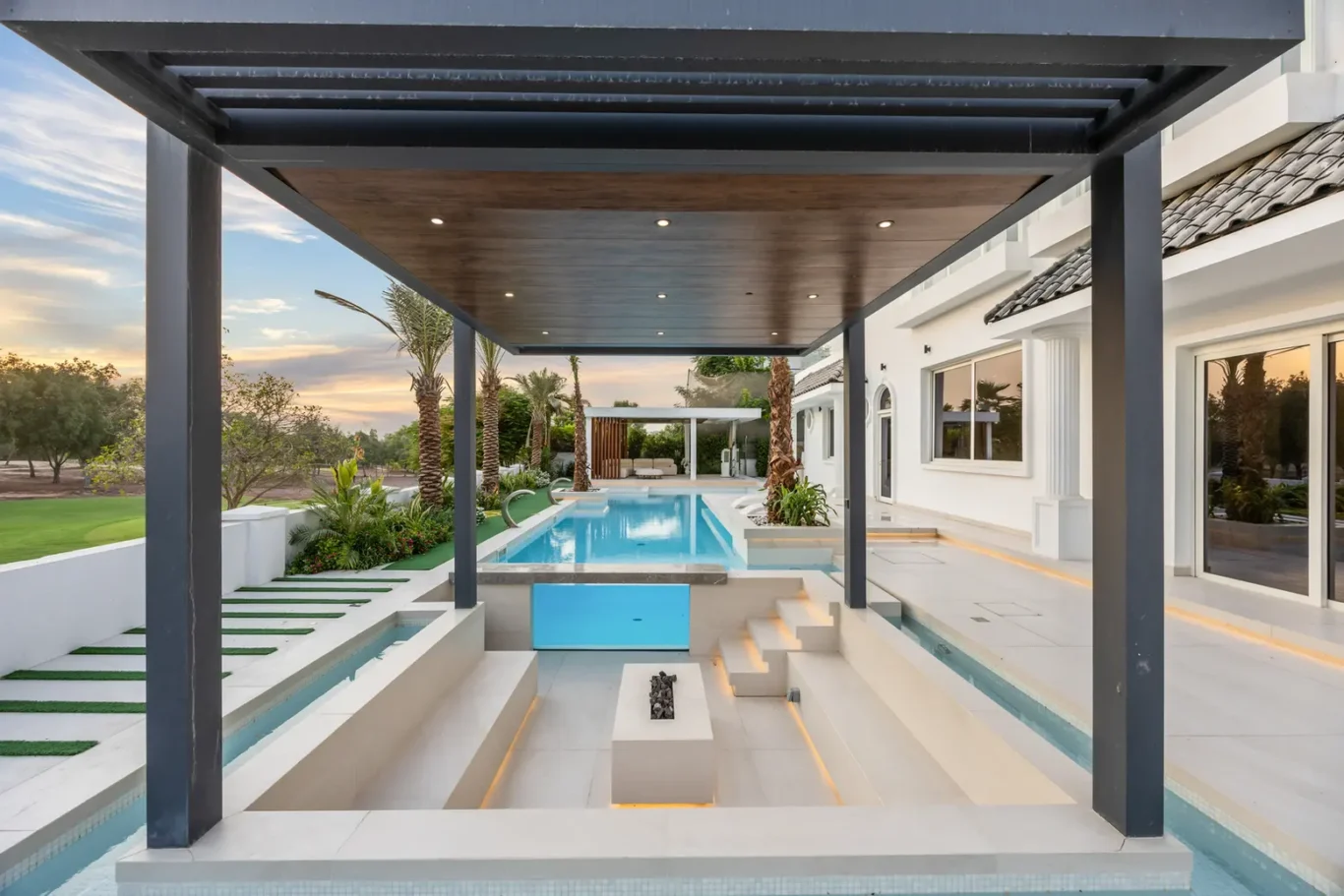 Modern backyard with a rectangular pool, white stone patio, and a dark-framed covered seating area alongside a white villa at sunset.