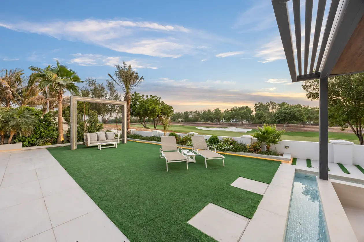 Modern outdoor patio with artificial turf, beige loungers, and a swing seat under a pergola, overlooking a green lawn area and golf course beyond.