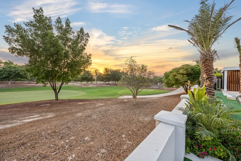 Sunset over a golf course with green fairways, trees, and tropical plants along a white railing in the foreground.