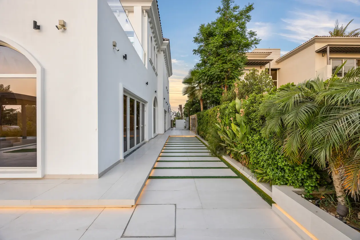 White modern building with glass doors along a paved walkway and a lush hedge on the right, during daytime.