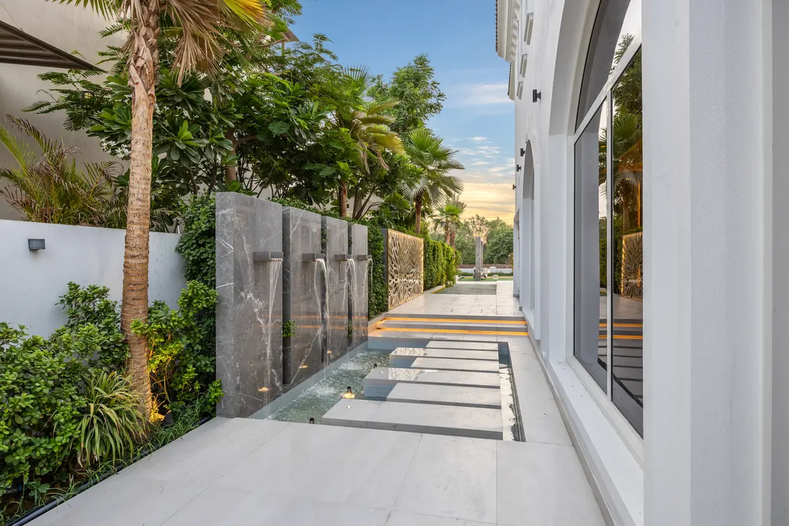 Modern outdoor courtyard with a water feature and vertical stone spouts along a low wall, flanked by tropical plants and a white building facade.
