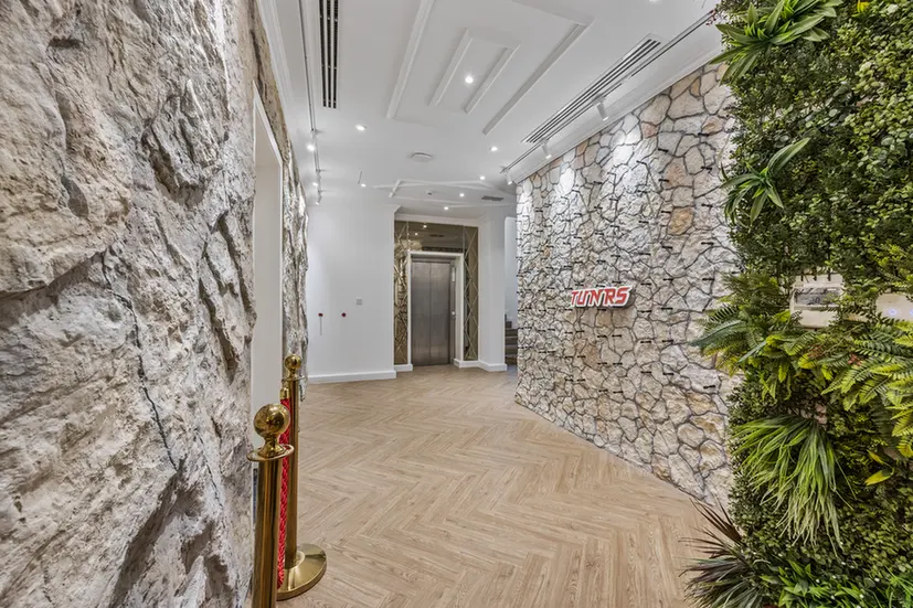 Modern lobby hallway with stone accent walls, herringbone wood floor, and an elevator at the far end; a vertical garden is on the right.