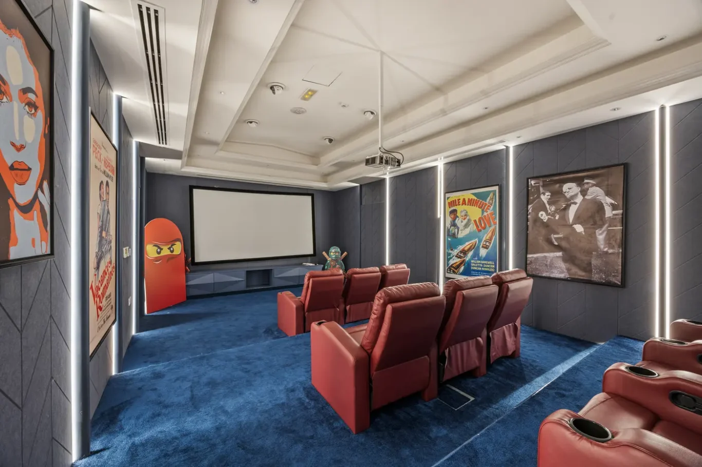 Home cinema with blue carpet, red leather recliner seats facing a large projection screen and movie posters on dark walls.