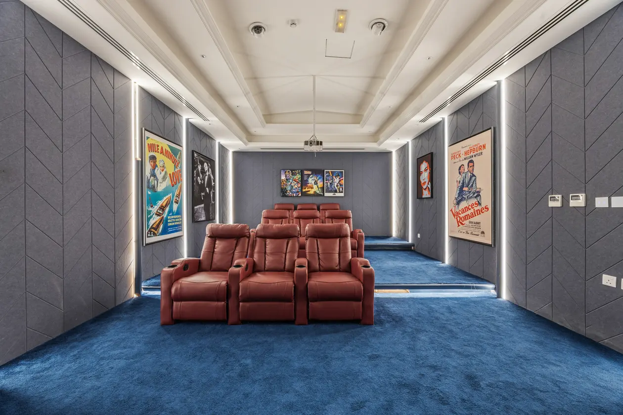 Home theater room with red leather recliner seats arranged in two rows and vintage movie posters on gray walls with blue carpeted floor.