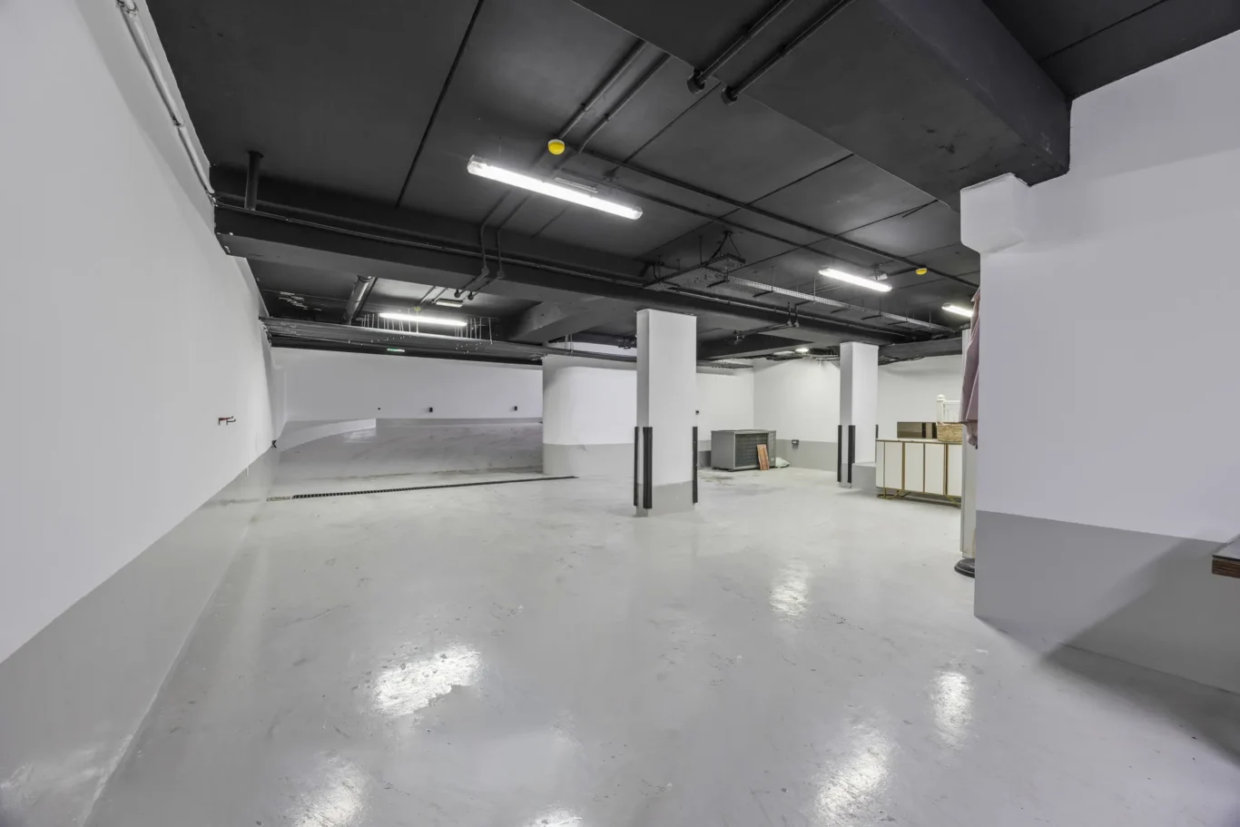 Empty basement parking area with white walls, gray concrete floor, and exposed dark ceiling pipes and fluorescent lights nearby.