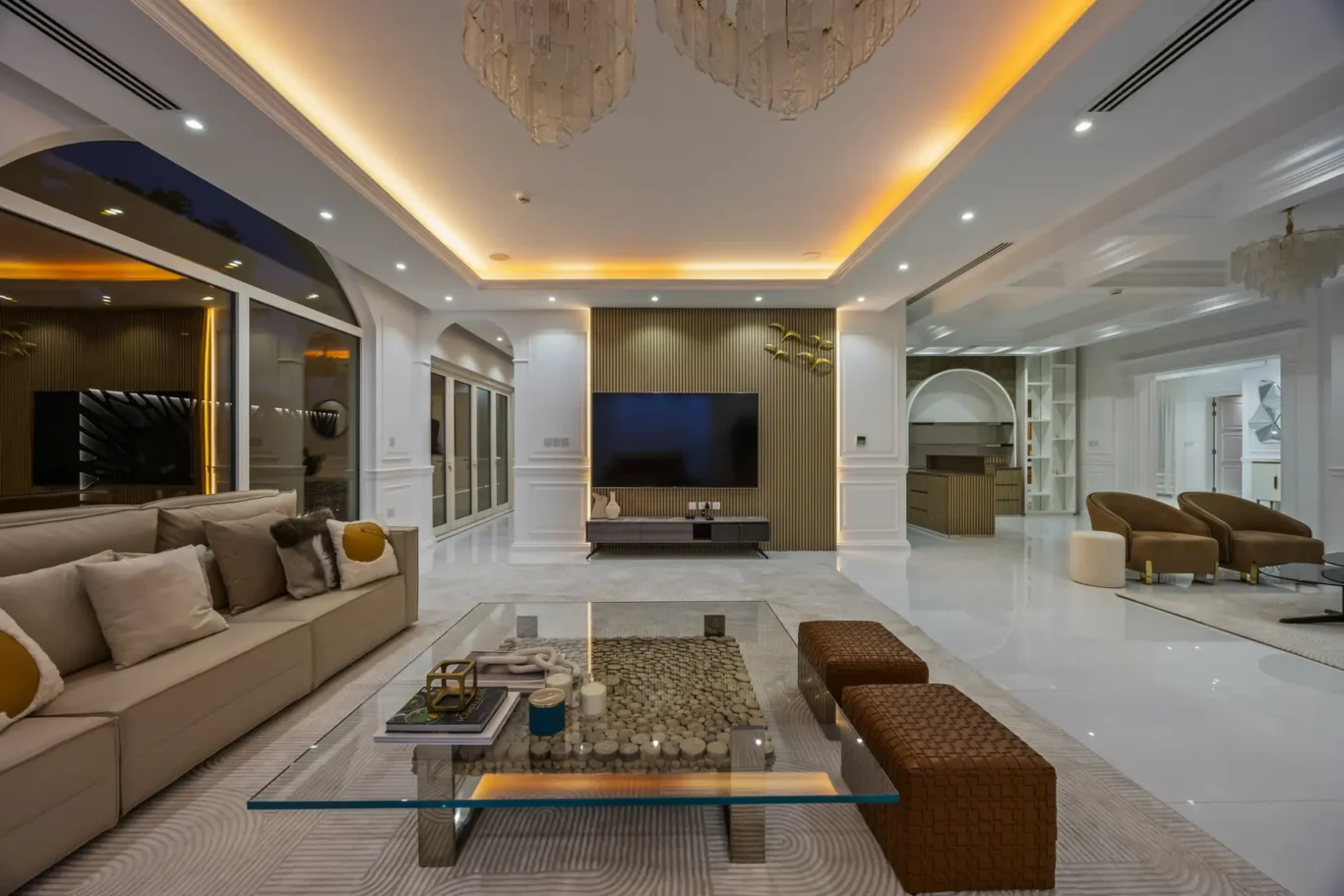 Luxurious open-plan living room with beige sofa, glass coffee table, and a wall-mounted TV on a gold-accented panel.