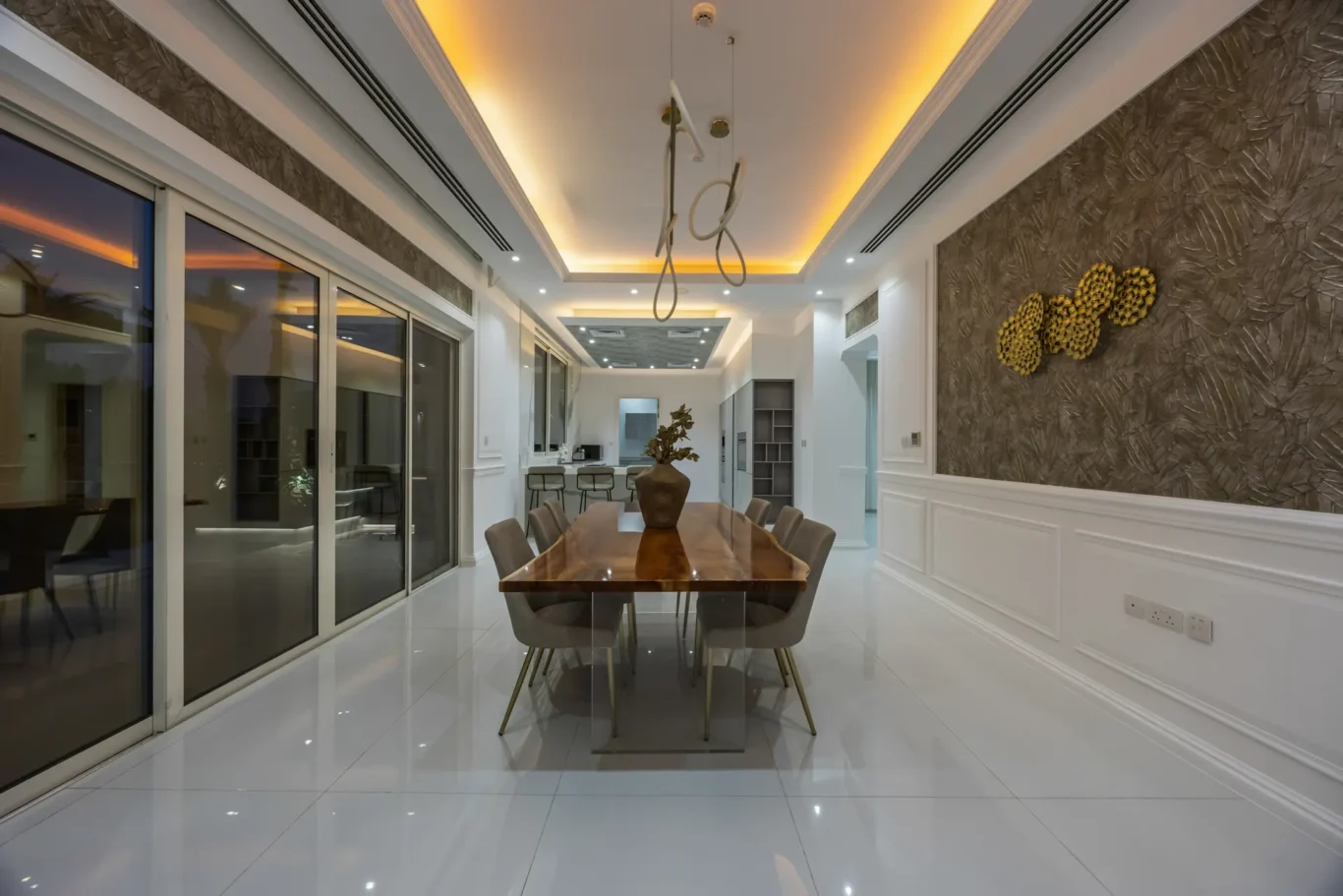 Modern dining room with a long wooden table and gray chairs, glass sliding doors, and textured wall art illuminated by warm ceiling lighting
