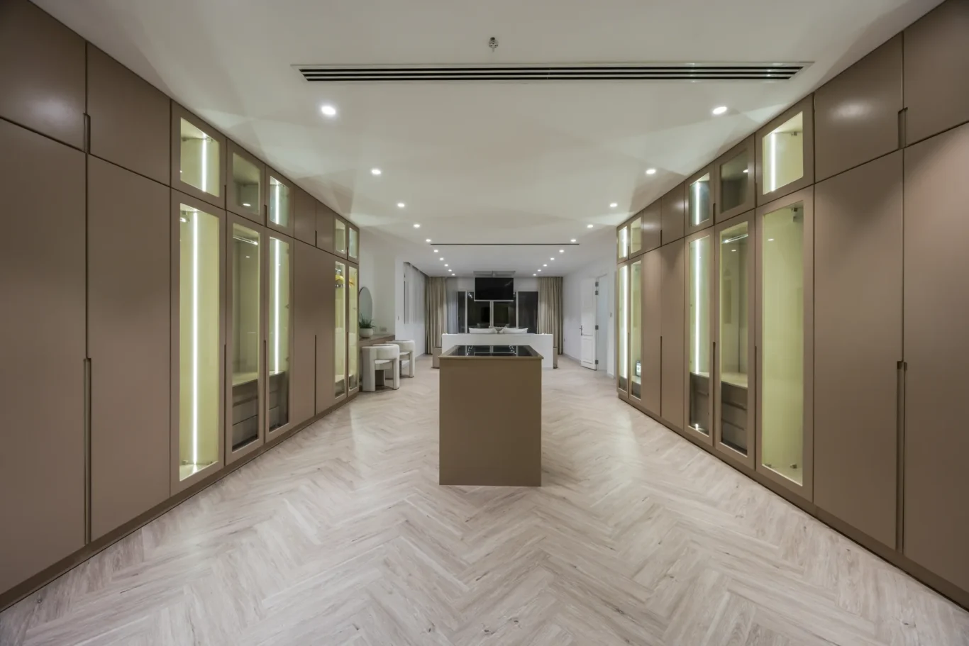 Walk-in closet with brown floor-to-ceiling cabinets, glass-front display cases, and a central island in a bright showroom.