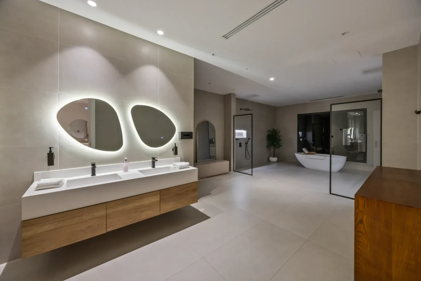 Modern bathroom showroom with double sink vanity, backlit oval mirrors, and glass-enclosed shower near a freestanding tub.
