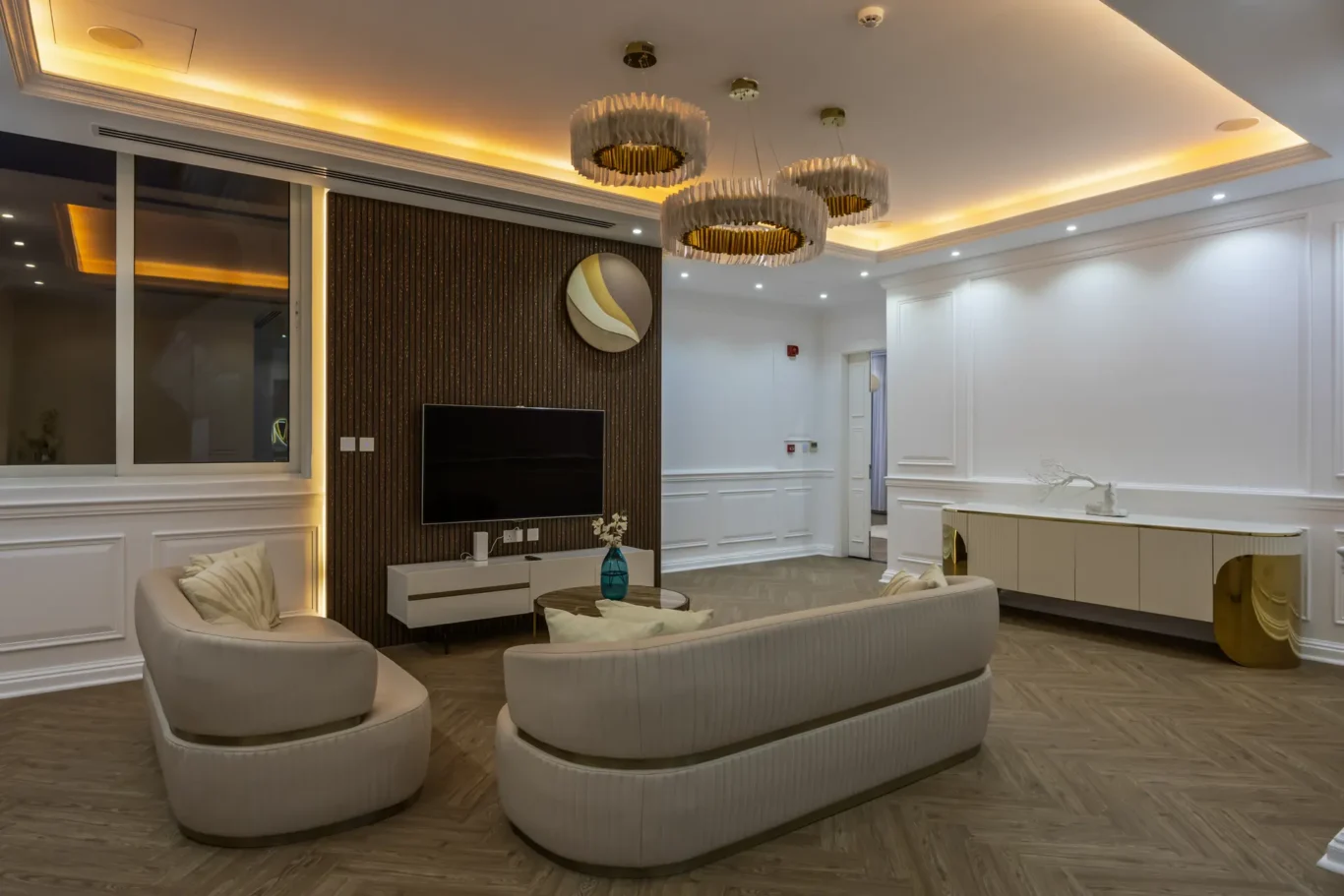 Luxurious living room with curved beige sofas, herringbone wood floor, and a dark wood TV wall with gold-accent chandelier clusters.