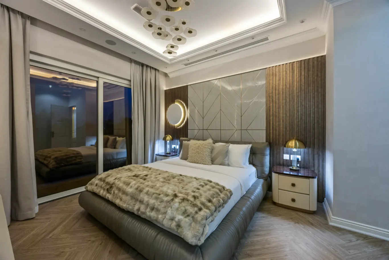 Luxurious hotel bedroom with a king bed, fur throw, padded headboard, and geometric wall panels, plus a round backlit mirror and nightstands.