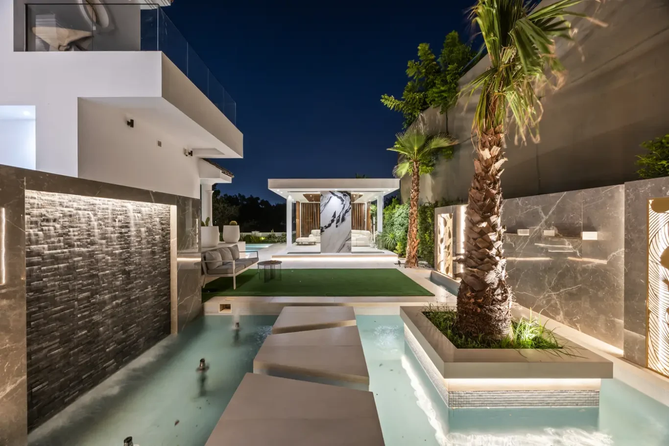 Modern nighttime backyard with a shallow pool, white stepping stones, palm trees, and contemporary seating.