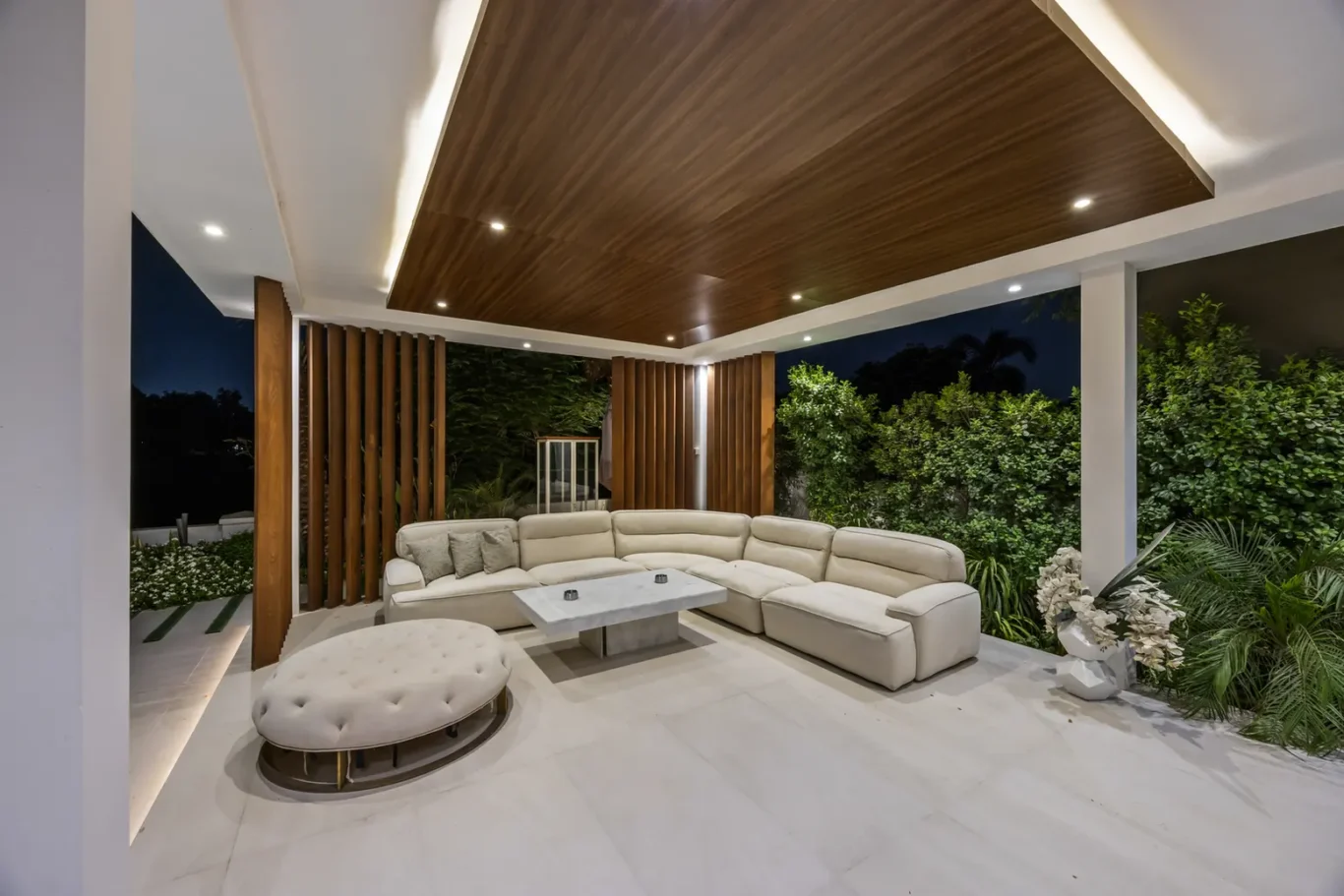 Outdoor covered patio with a large beige sectional sofa, round tufted ottoman, and a square marble coffee table under a wooden paneled ceiling at night.