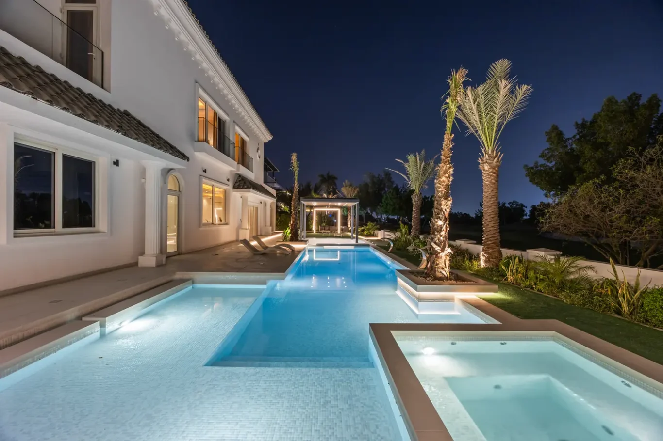 Luxury white villa at night with a long, illuminated swimming pool and palm trees along the patio area