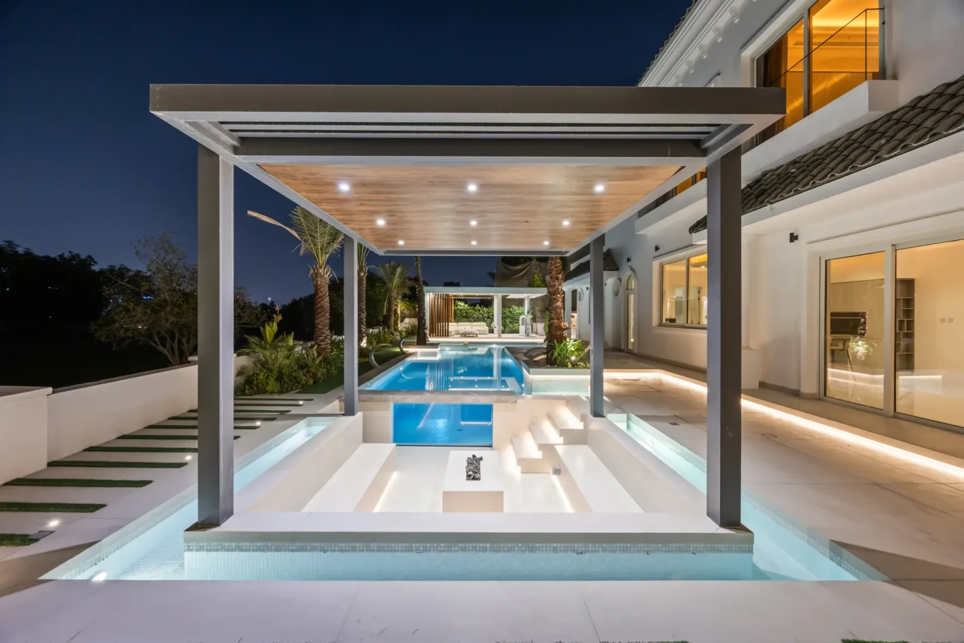Night view of a modern villa with an illuminated rectangular pool, white steps, and a wooden pergola over the terrace.