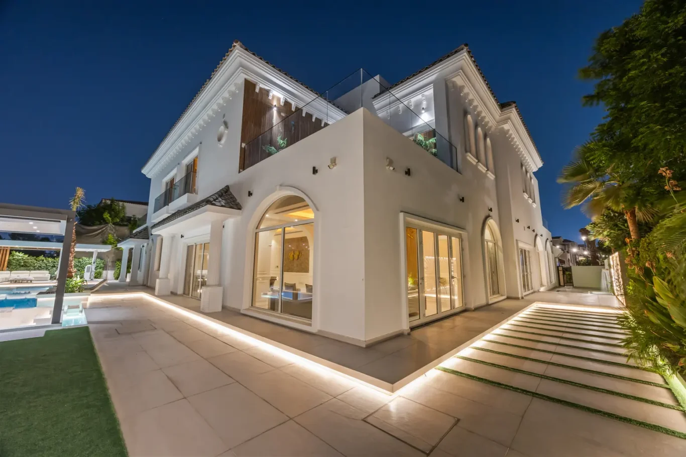 Modern white villa at night with glass balcony, arched windows, and warm patio lighting by a pool area.
