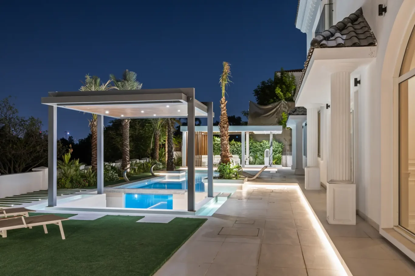 Luxurious backyard at night with a illuminated rectangular pool, white modern architecture, and palm trees surrounding a covered seating area.