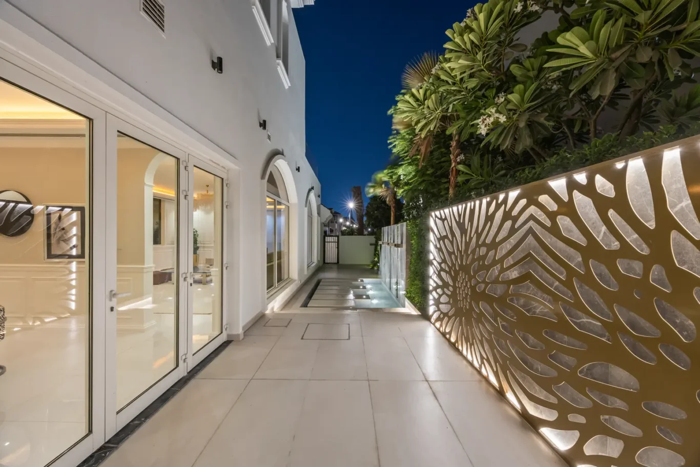 Nighttime patio scene: glass doors to a white house on the left, decorative illuminated metal screen on the right, and a lit path with a slender pool ahead.