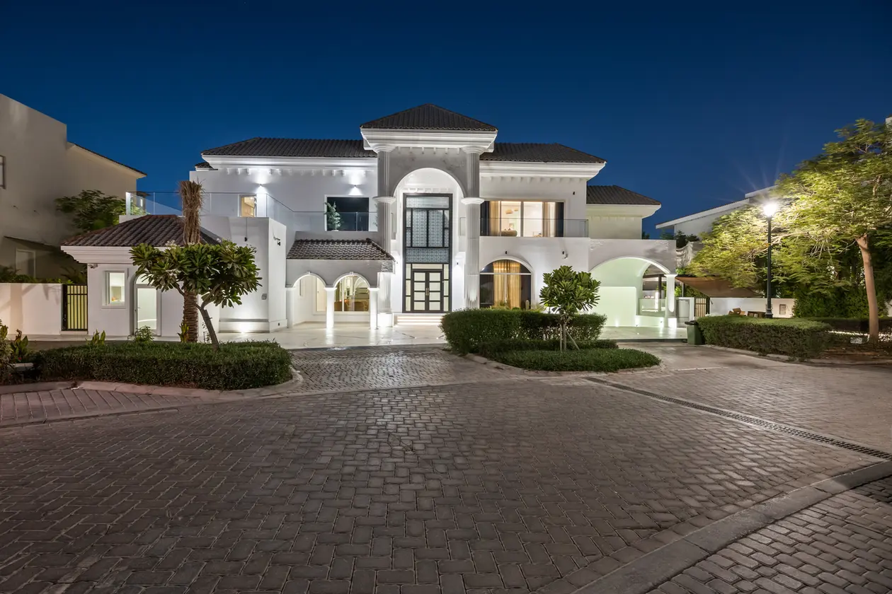 White modern mansion at night with a well-lit entrance and cobblestone driveway.
