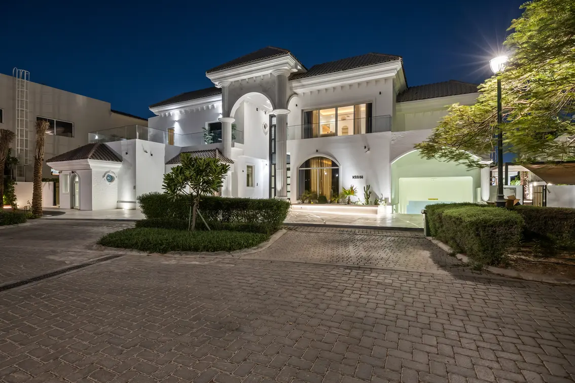White Mediterranean-style villa at night with a grand arched entry, columns, and warm interior lights.