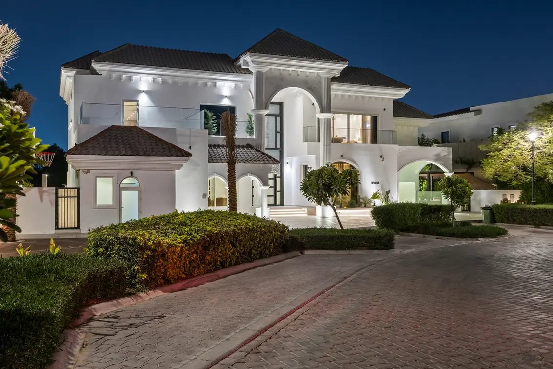 White Mediterranean villa at night with an illuminated arched entrance and manicured landscaping in front of a cobblestone driveway