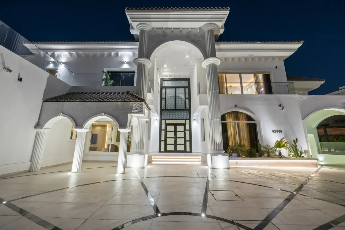 Elegant white mansion at night with a grand entrance, two fluted columns, and arched doorway leading to illuminated doors in a modern courtyard.