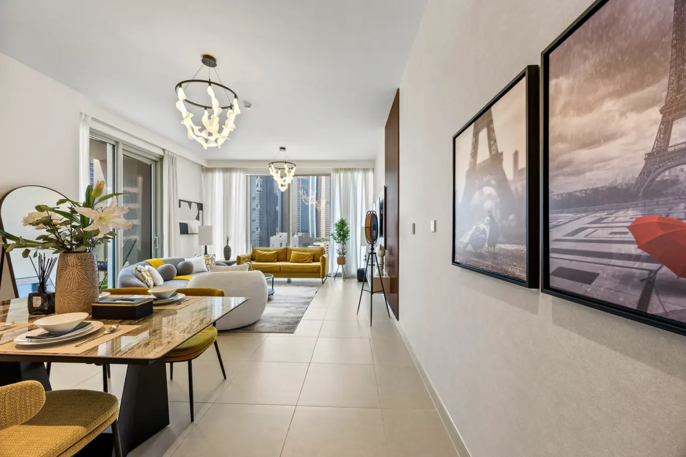 Bright open-plan living-dining room with a mustard sofa, marble dining table, and white seating; large windows show a city skyline.