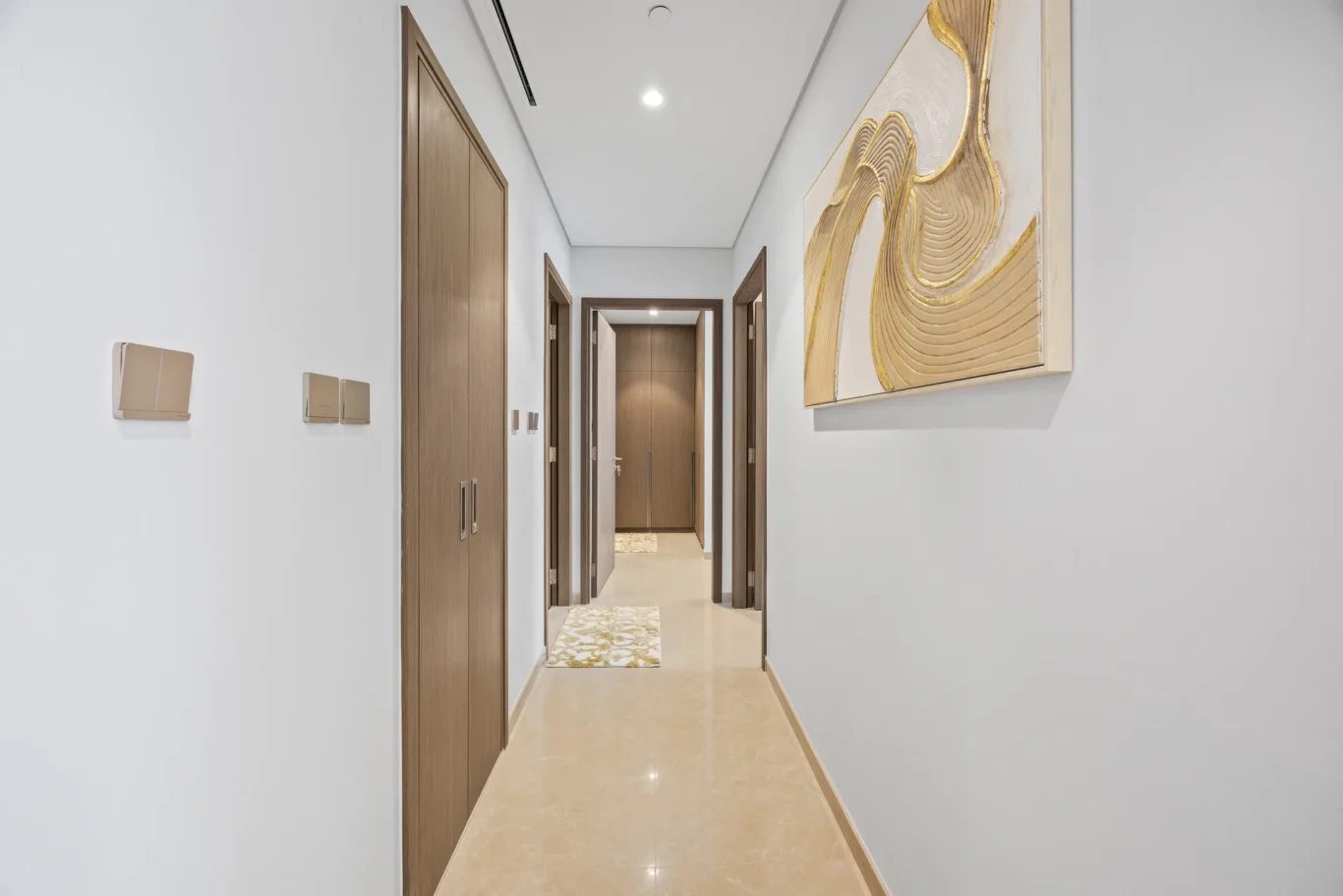 Narrow hallway with wooden doors on both sides, beige tiled floor, and a large abstract gold-accent painting on the right wall.