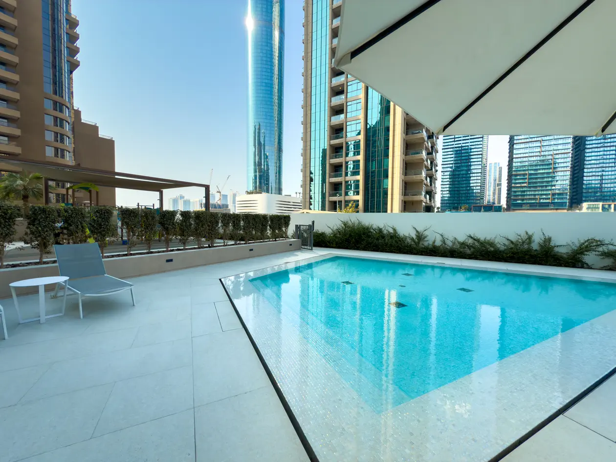 Rooftop pool with blue water, white tile deck, a single gray lounge chair and small round table, with tall glass buildings in the background.