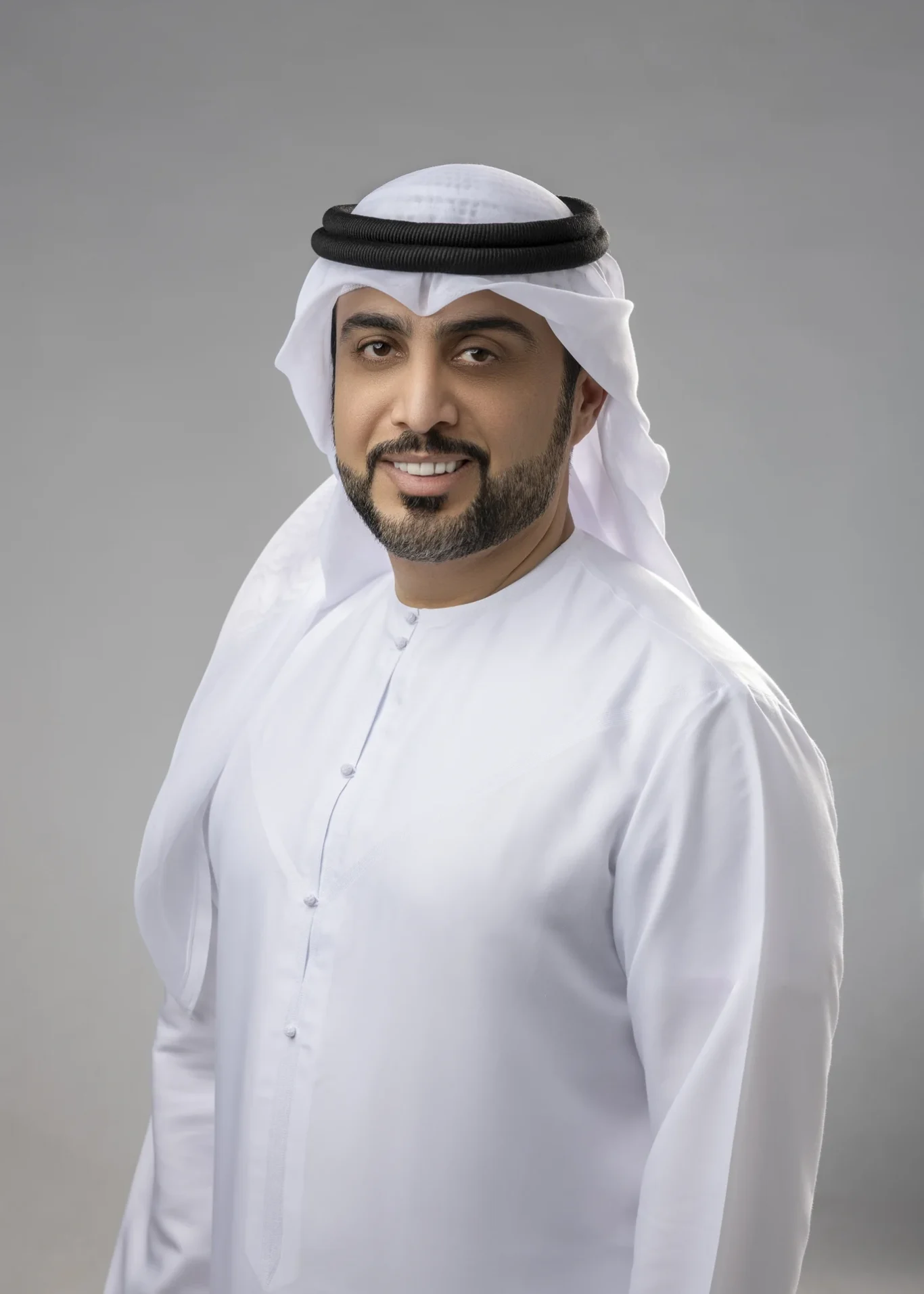 Smiling man in a white kandura and black agal against a gray studio backdrop.