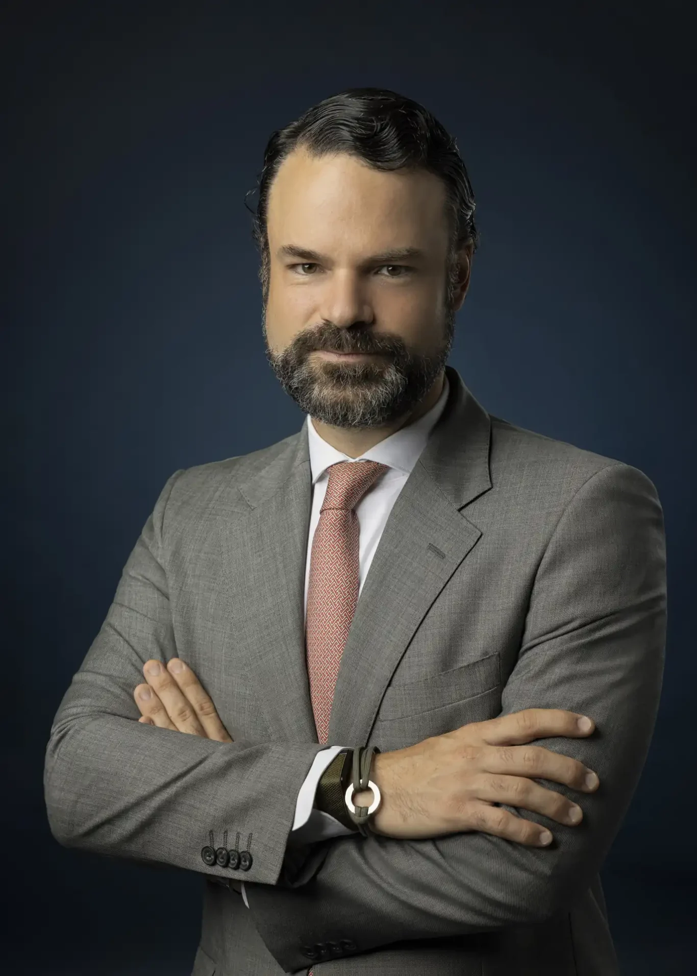 Confident businessman portrait with arms crossed in a gray suit and red tie against a dark background.