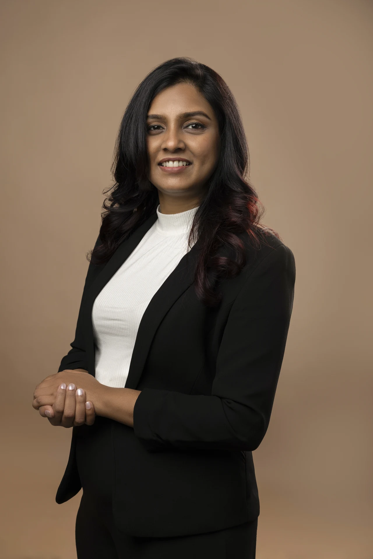 Professional woman in a black blazer and white top, standing with hands clasped in front against a neutral tan backdrop.