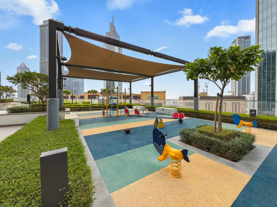 Rooftop playground with tan shade sails, colorful rubber flooring, and city skyscrapers in the background.