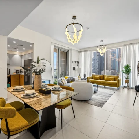 Open-concept living and dining area with mustard chairs, marble table, gray sofa, and large windows showing city skyline on a bright day.