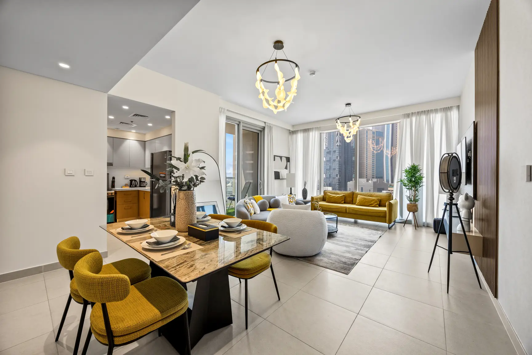 Open-concept living and dining area with mustard chairs, marble table, gray sofa, and large windows showing city skyline on a bright day.