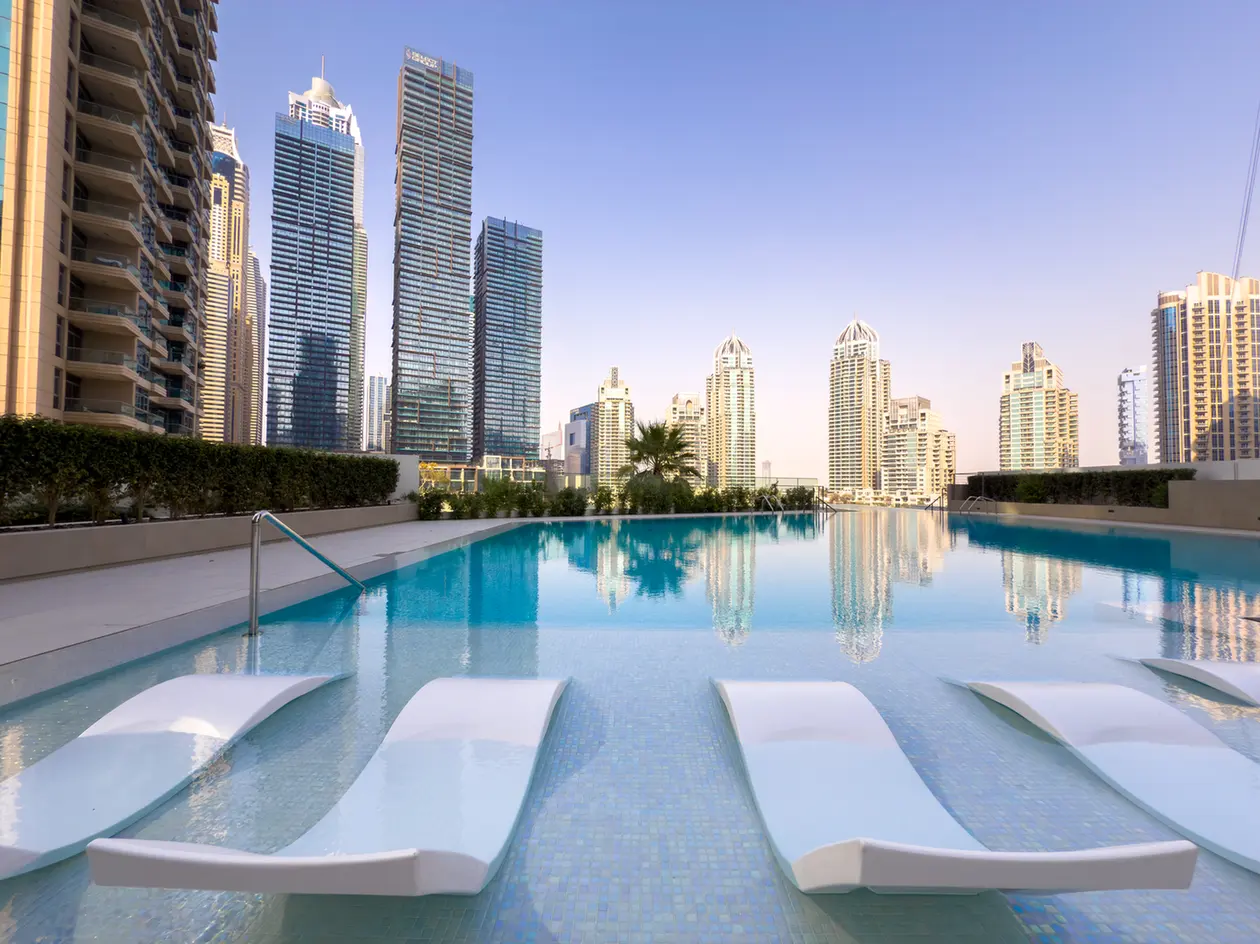 Rooftop pool with white lounge chairs and a city skyline of tall glass buildings in the background.