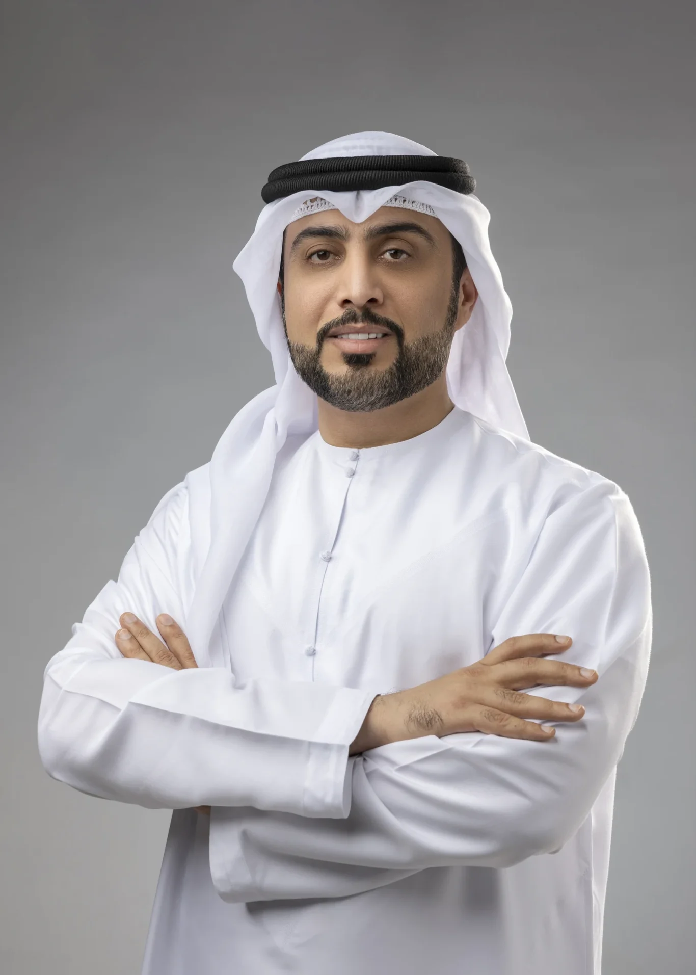 Portrait of a man in traditional Emirati dress (white kandura and ghutra with agal), arms crossed, facing the camera against a gray studio background.