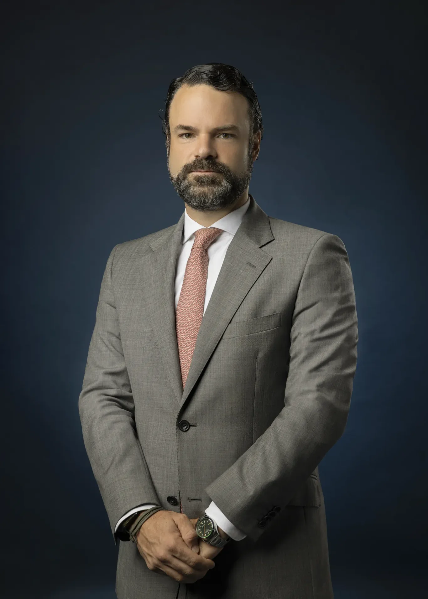 Portrait of a man in a gray suit with a pink tie, standing with hands clasped in front, against a dark blue studio backdrop.