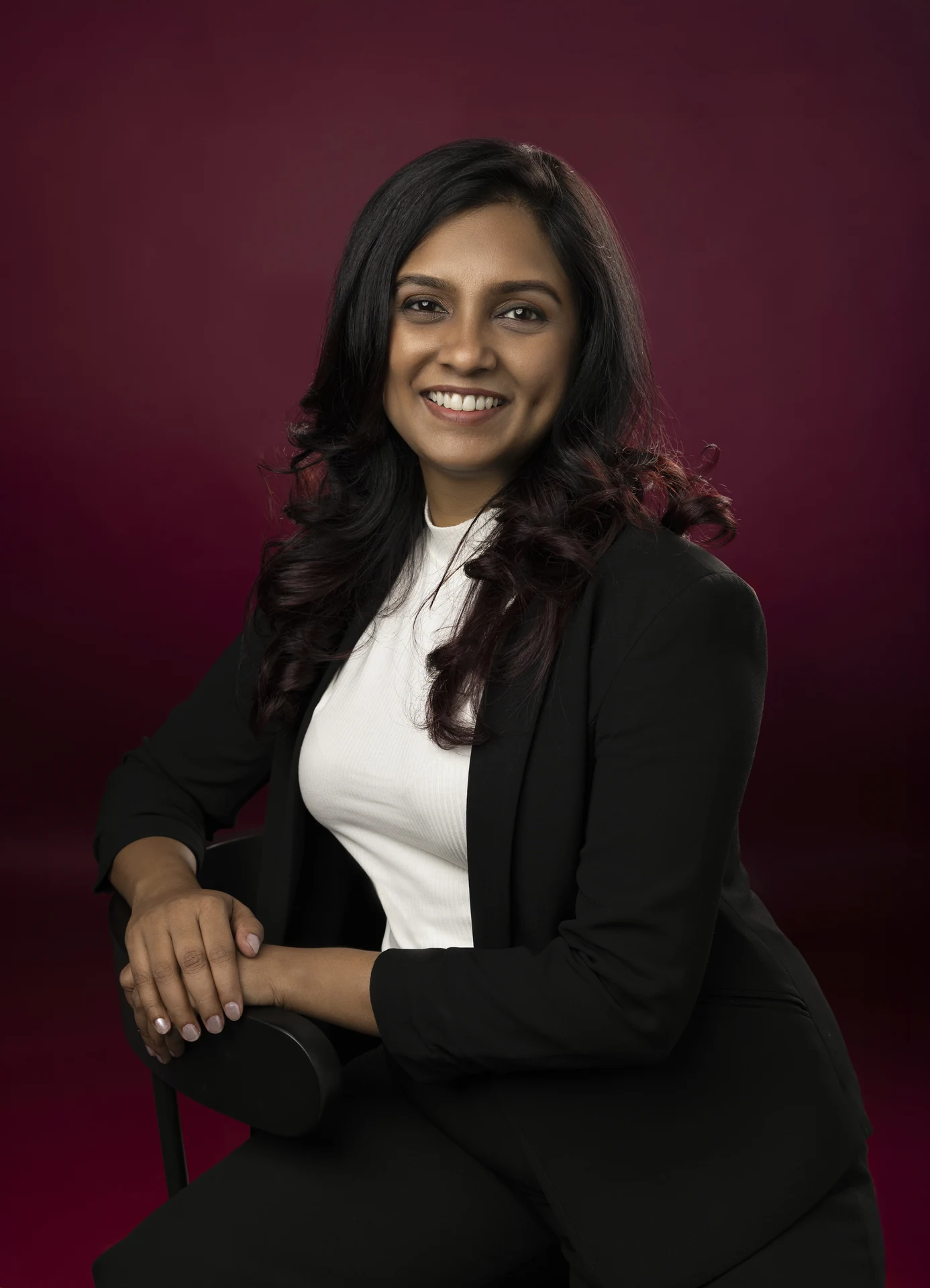 Professional woman with dark hair, wearing a white blouse and black blazer, seated and smiling against a burgundy backdrop.