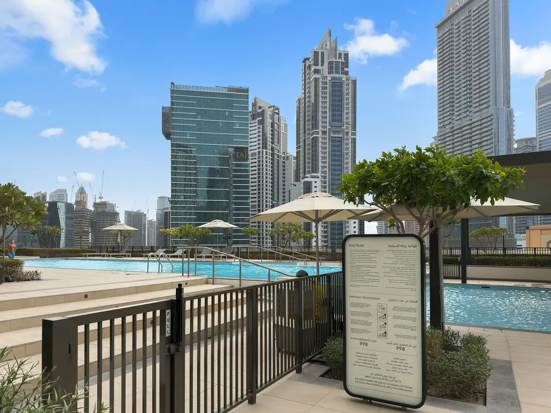 Rooftop pool area with lounge umbrellas and a city skyline of tall glass buildings in the background under a blue sky