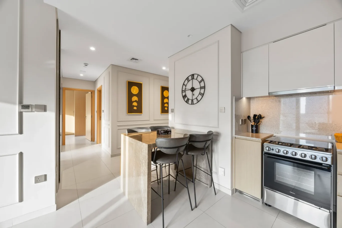 Modern kitchen with marble island, four black bar stools, white cabinets, and a large wall clock.