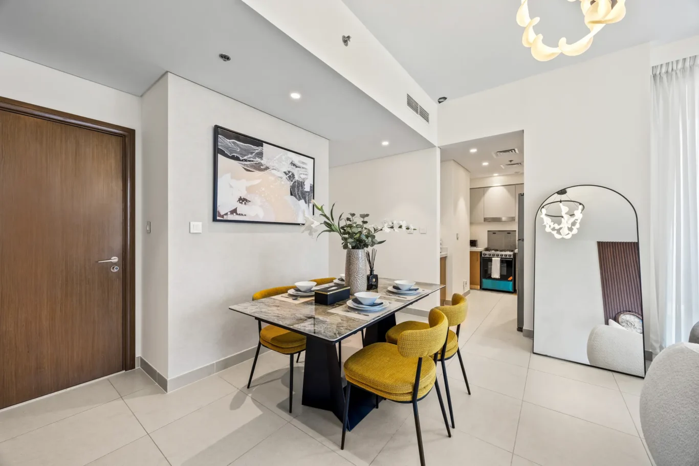 Open-plan dining area with a marble table, six mustard chairs, white walls, and a large round mirror nearby.