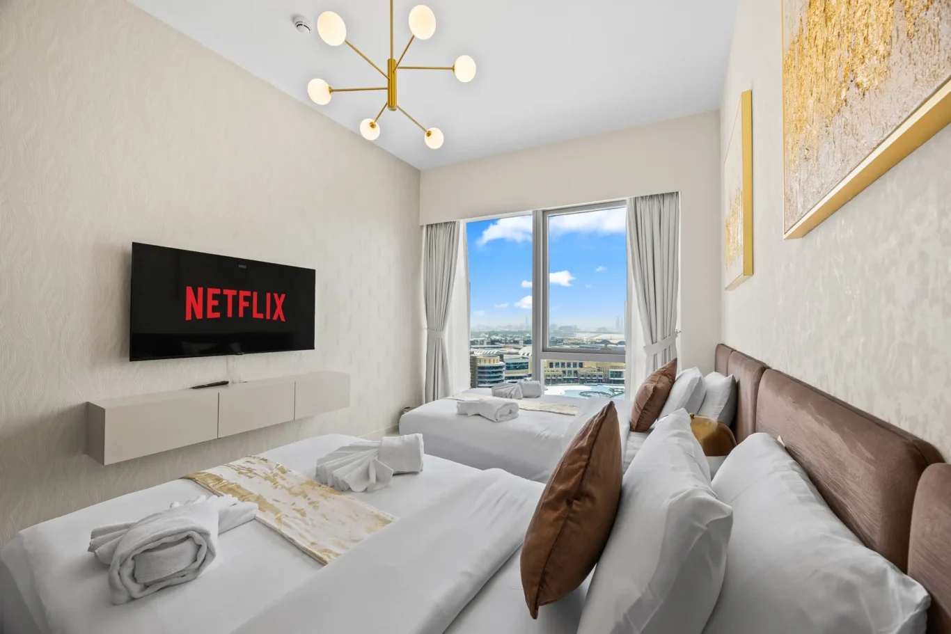 Contemporary hotel room with two white beds, brown throw pillows, a wall-mounted Netflix screen, and a large window offering a city view.